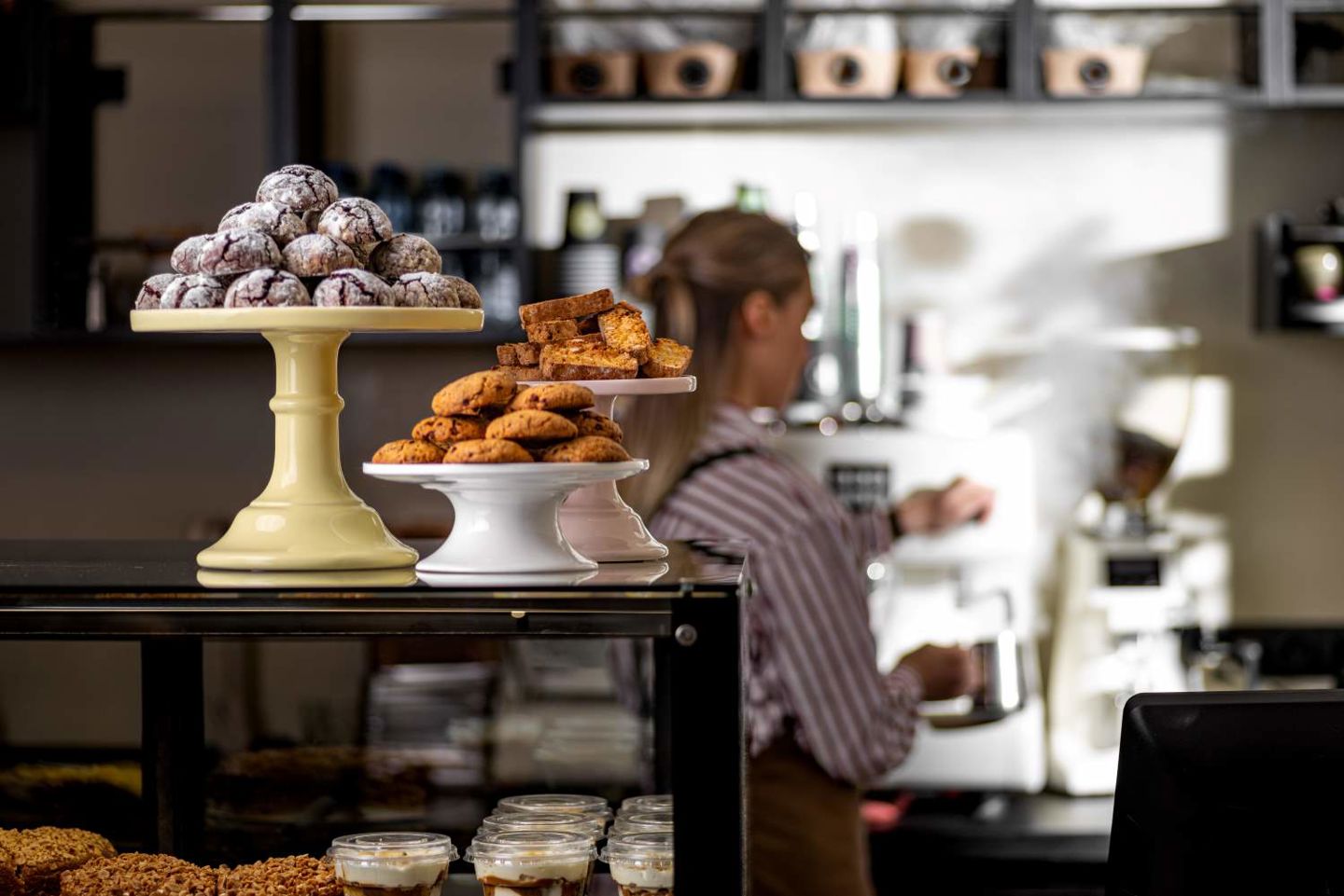 Bilde av en ung kvinne som jobber i en kafé, med kaker og kjeks på disken 