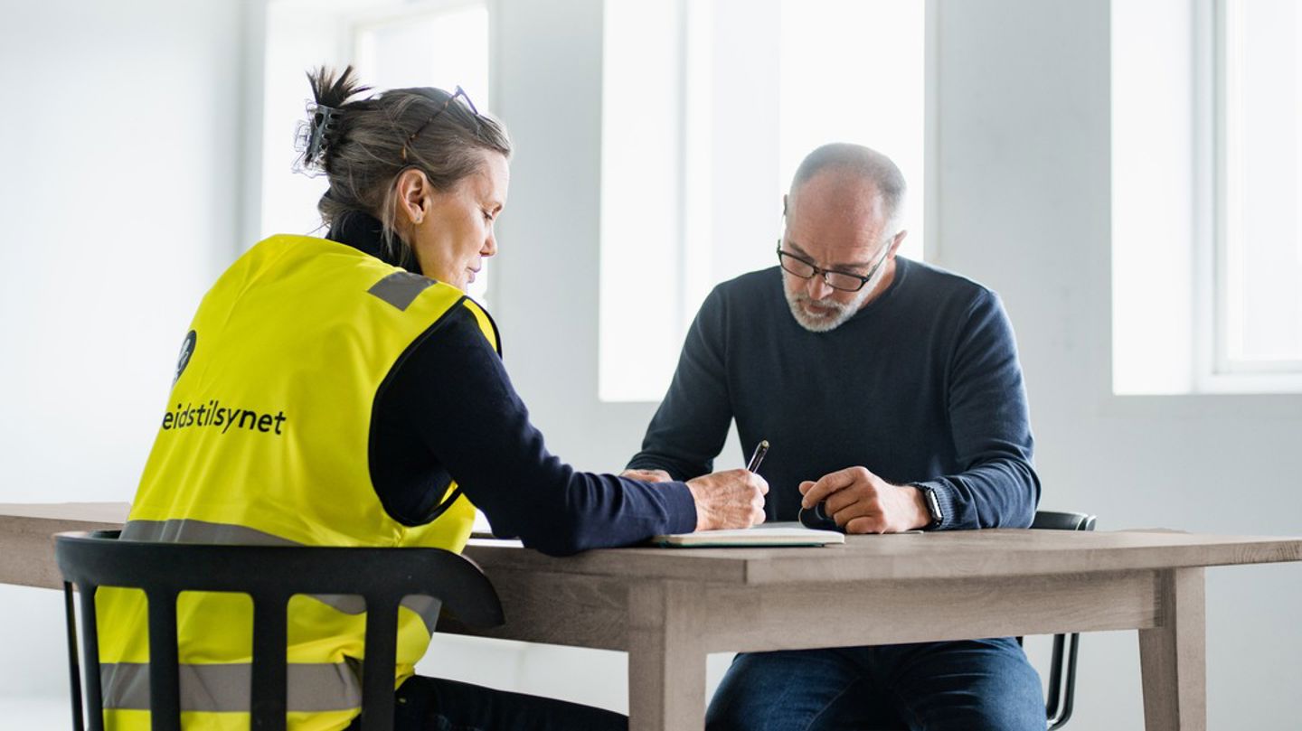 Bildet viser to personer som sitter ved et bord. Den ene har på seg gul Arbeidstilsynet-vest. 
