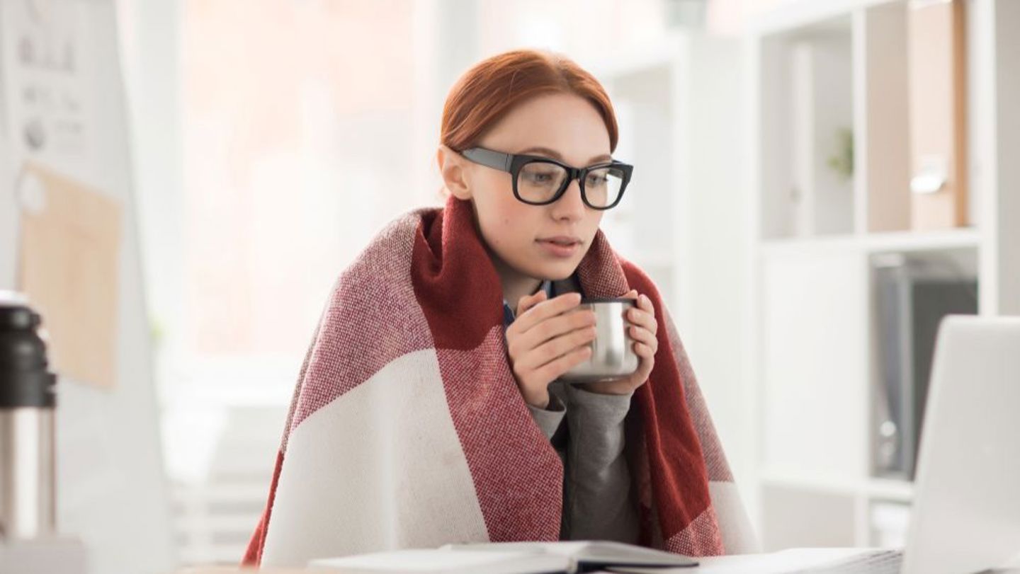 Bildet viser ei dame som sitter på en kontorarbeidsplass med pledd på seg og en kopp i hendene. 