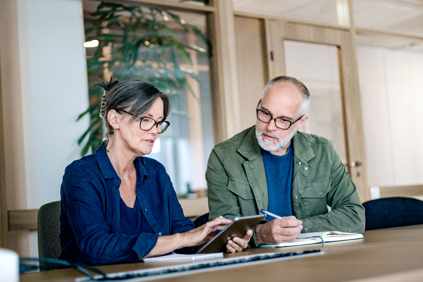 Kvinne og mann sitter på e kontor, kvinnen ser på en ipad og mannen noterer i ei bok