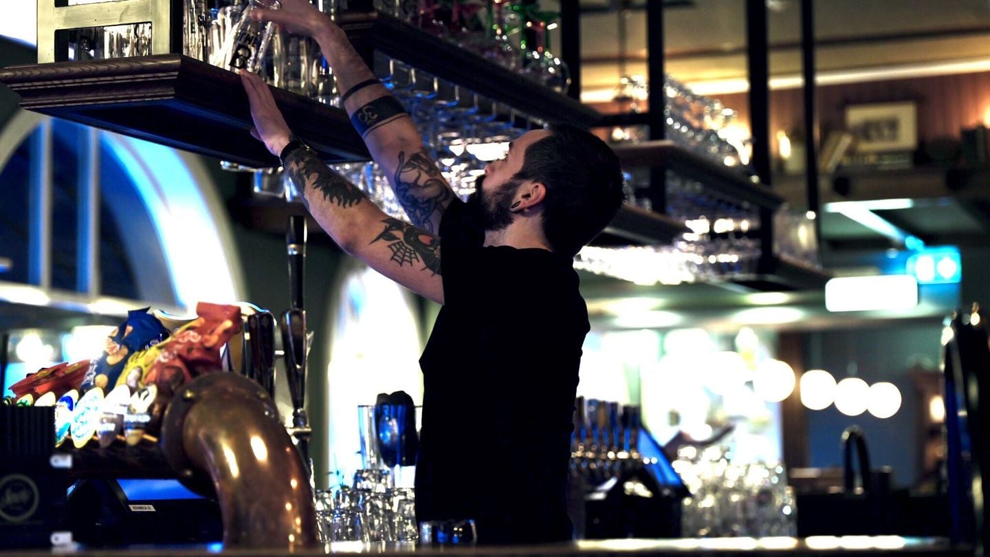 En mann står bak en bar og tar glass ned fra et stativ. 