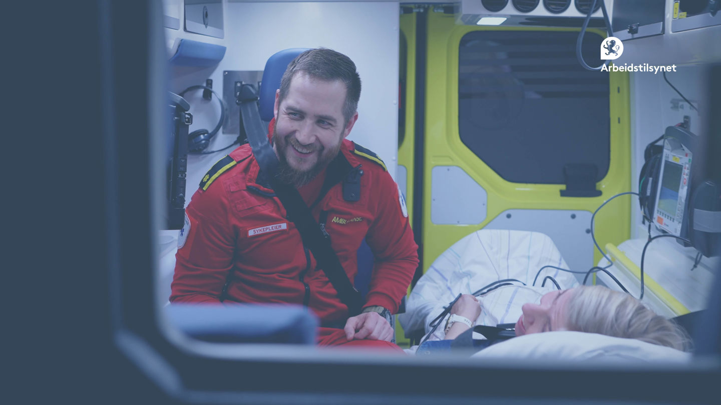 Bilde fra innslag fra sykehus i webinaret. Det er tatt bakover i en ambulanse. Ambulansemedarbeideren sitter og snakker med en pasient som ligger i senga. Han smiler.