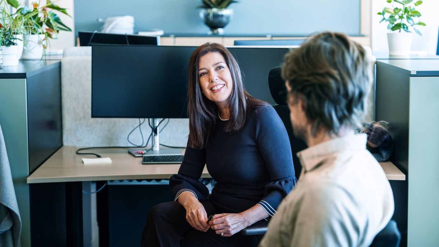 Bildet viser en kvinne på en kontorarbeidsplass i dialog med kollega.