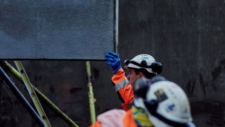 Bildet viser to personer på en byggeplass. Den ene av dem tar imot ei plate som senkes med ei kran.