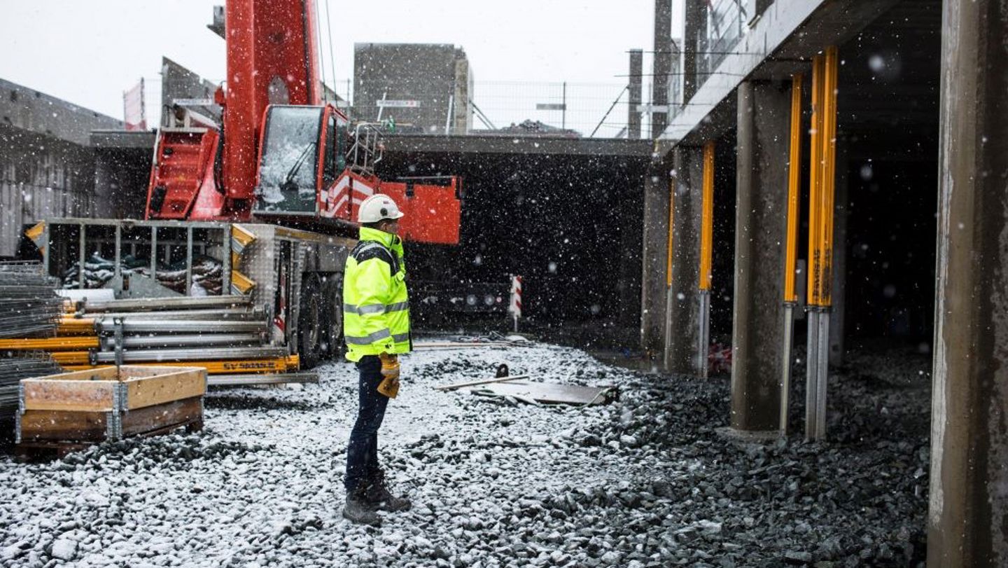 Bildet viser en mann med gul jakke som står på en byggeplass i snøvær.