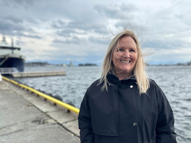 Kvinne står i vinden på ei brygge med sjø og en båt i bakgrunnen 