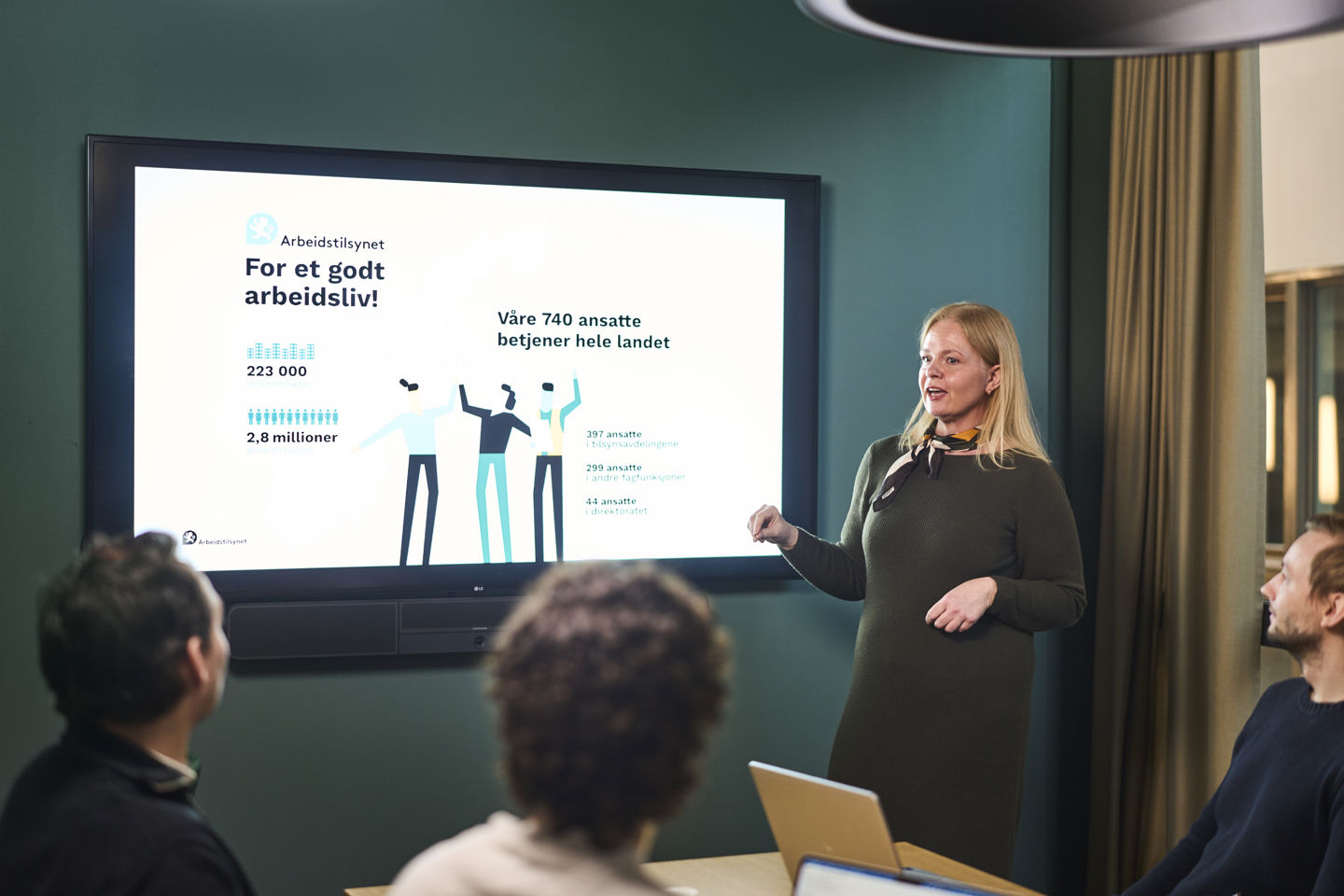 Ei dame står foran en skjerm med en presentasjon med fakta om Arbeidstilsynet. Vi ser tre tilhørere bakfra. 