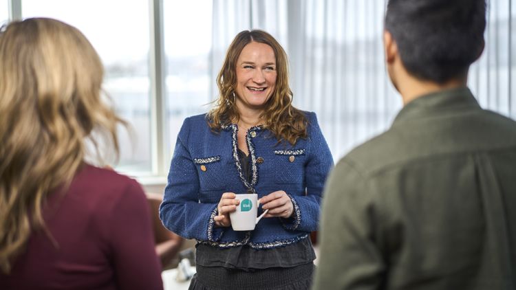 Dame med brunt langt hår står smilende med en kopp kaffe i hånda mens to mennsker vises bakfra