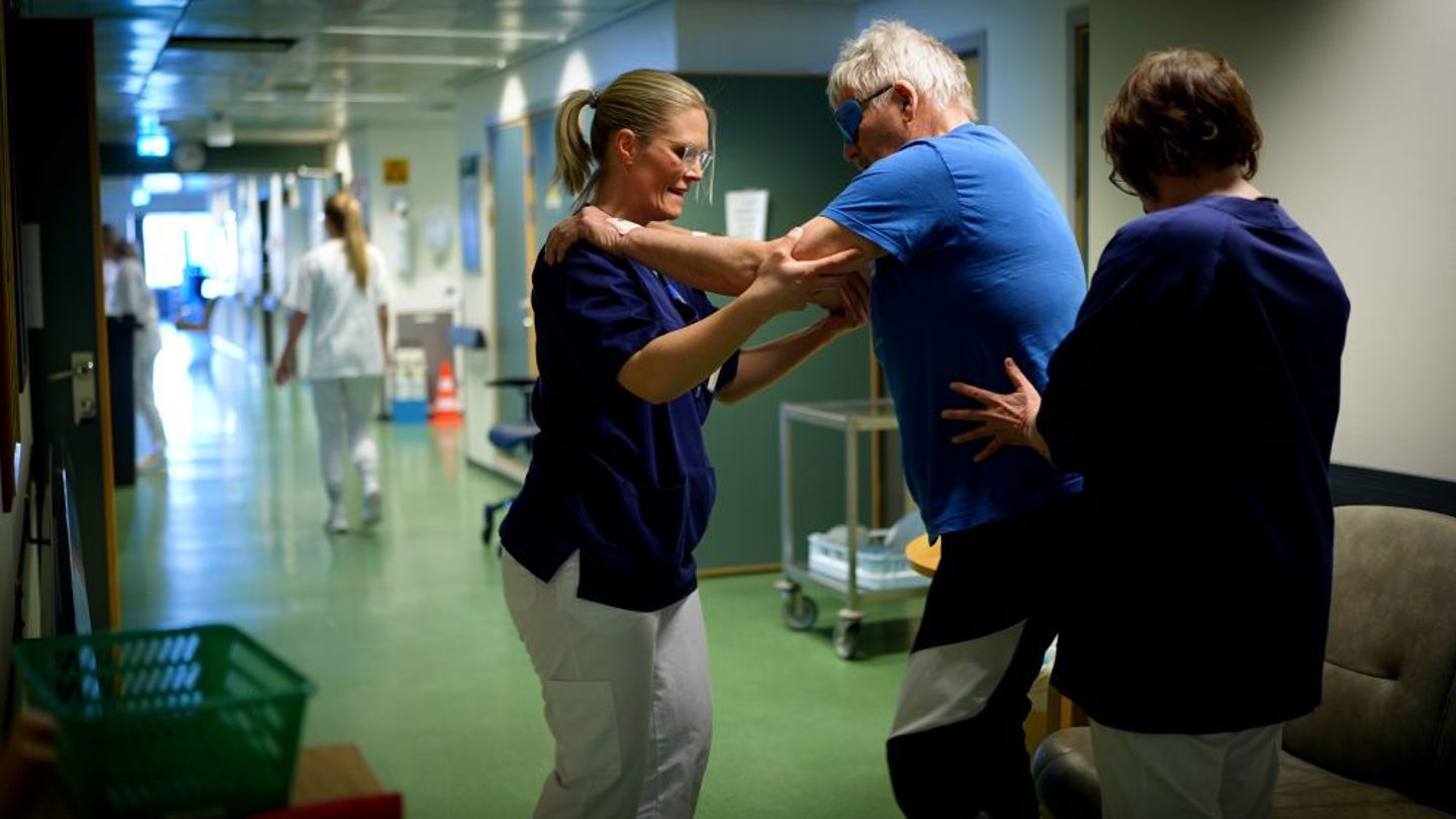 Bildet viser to fysioterapeuter som trener med en pasient i en sykehuskorridor.